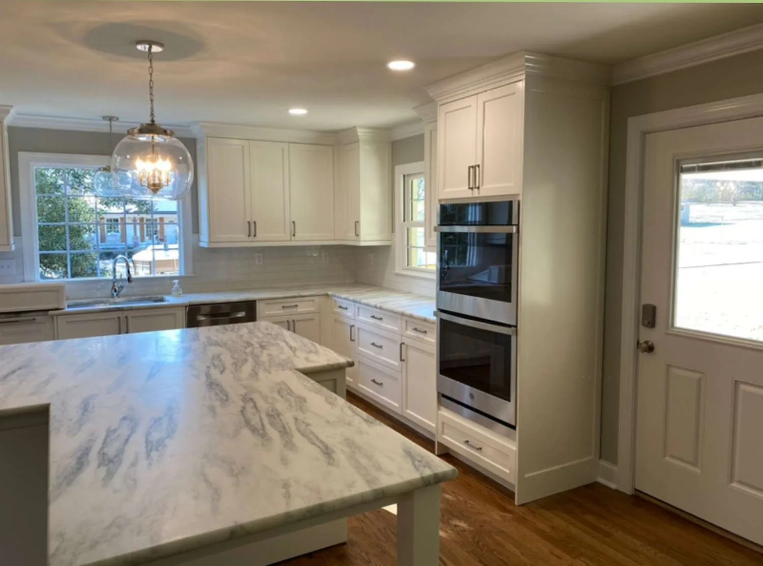 The same kitchen after renovation, with white cabinets, a marble island and a glass pendant